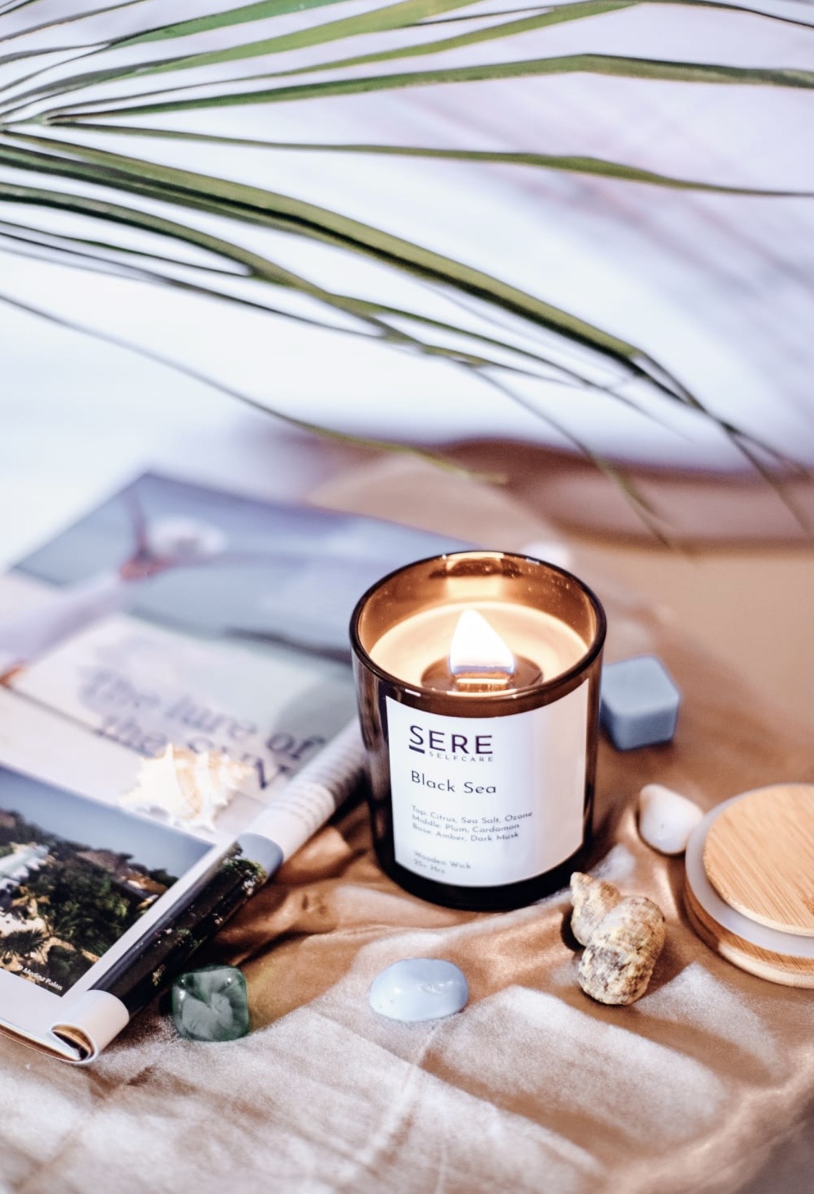 A lit Sere candle resting on draped fabric with palm fronds